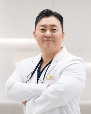 Doctor Ahn from Yonsei Bolt Dental Clinic, in a white lab coat with his arms crossed, standing in a bright, clean indoor area of the clinic. 