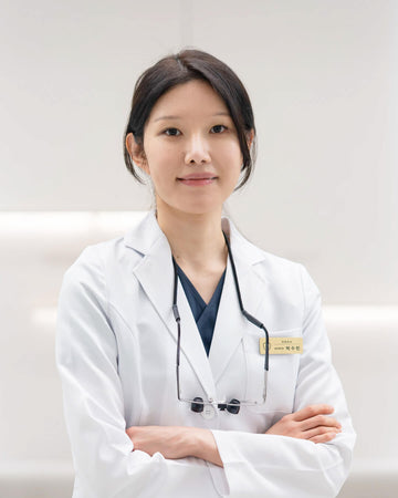 Doctor Park from Yonsei Bolt Dental Clinic, wearing a white medical lab coat over navy blue scrubs, with glasses around her neck and a name tag on her chest, standing with her arms crossed against a bright white background.